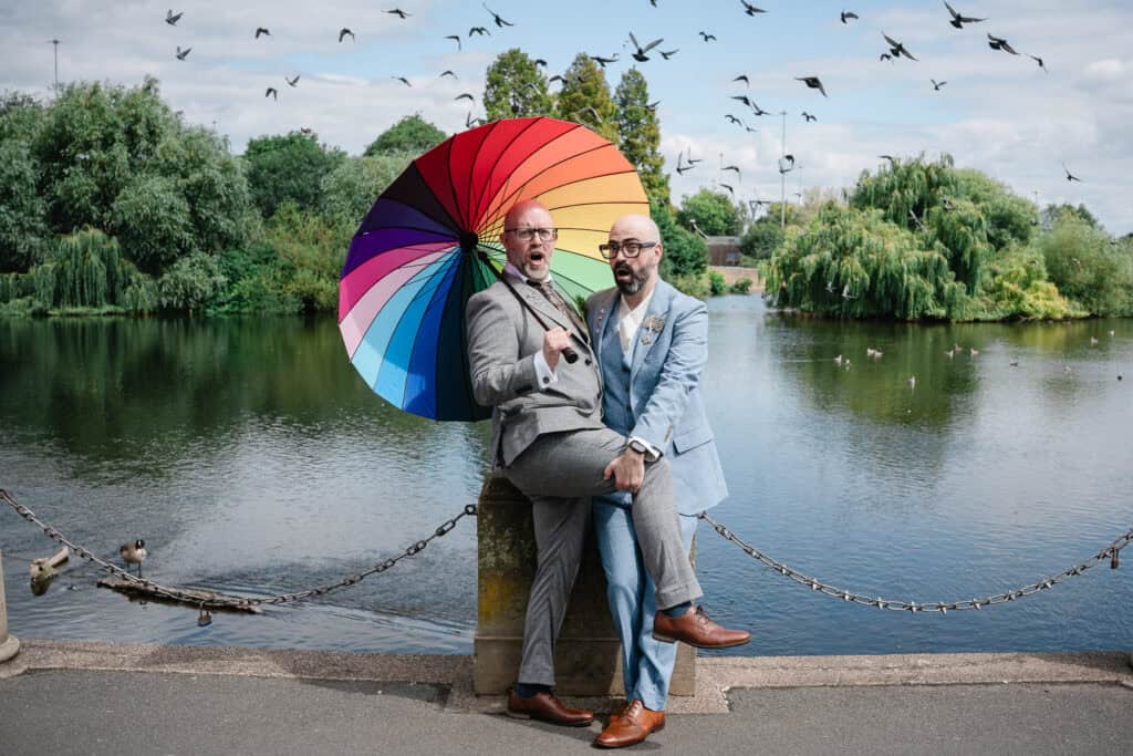 two grooms pose by the river in derby under a rainbow umbrella